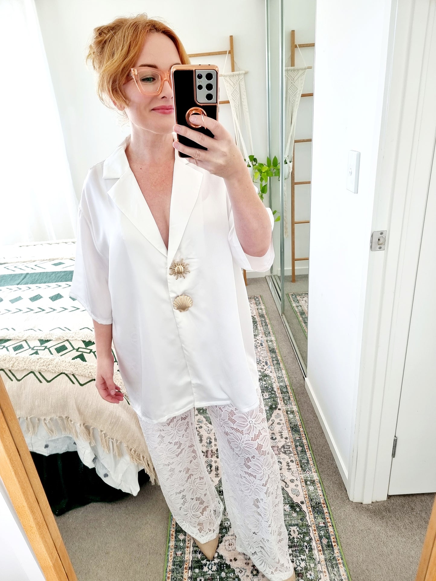 Tegan wearing the Amara Set in White by By Frankie. Oversized white satin shirt with gold shell and sun buttons, styled with sheer white floral lace pants. Tegan is an AU size 10 and wears the Medium.