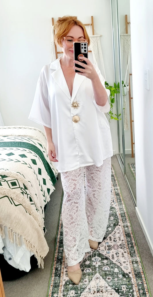 Tegan wearing the Amara Set in White by By Frankie. Oversized white satin shirt with gold shell and sun buttons, styled with sheer white floral lace pants. Tegan is an AU size 10 and wears the Medium.