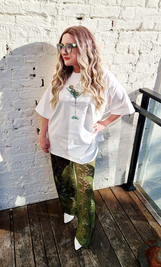 Tegan wears the Dirty Martini Set from By Frankie, featuring an oversized white t-shirt with a martini glass graphic and sheer moss green lace pants. She is standing on a timber deck against a white brick wall, styled with loose waves, statement green glasses, and white pointed-toe heels.